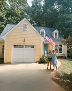 Homeowner standing in front of a yellow house holding a “Sold” sign, celebrating a successful home sale.